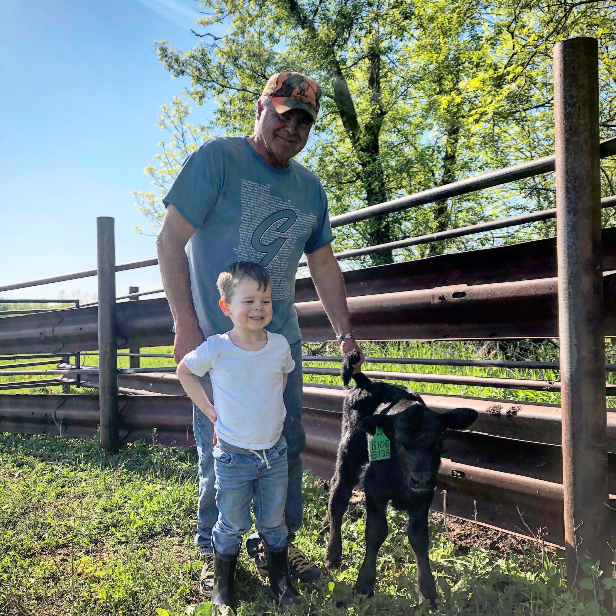 Tim Pettit stands behind his grandson Aidan beside a small black cattle and a metal fence.