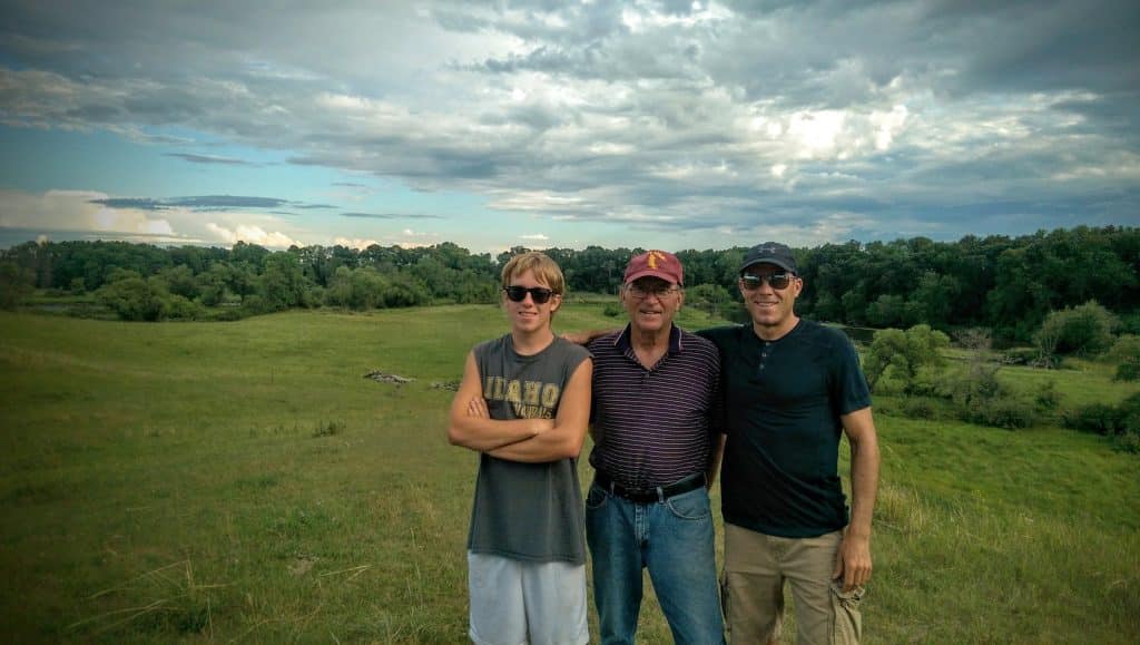 David Lindig standing in an open field with his arms around his grandson Ryan on the left and son Darin on the right, all smiling.