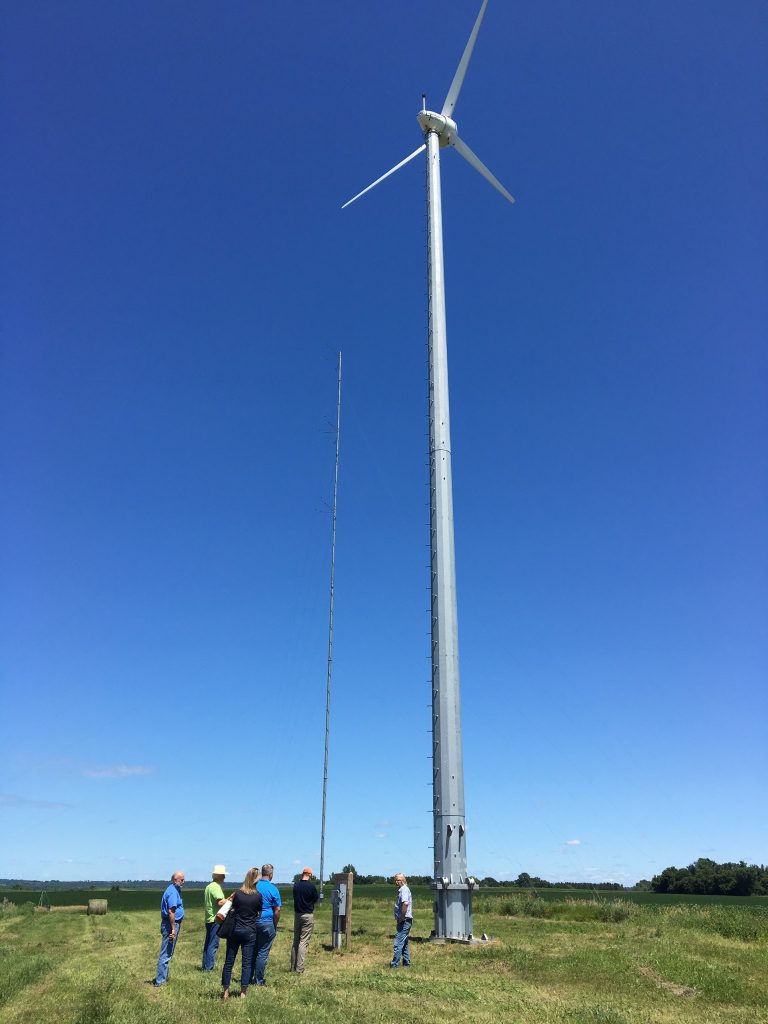 People gathered on a lawn underneath a tall wind turbine against a clear blue sky.